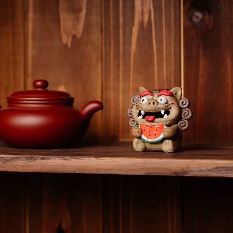 A handcrafted ceramic Pixiu backflow incense burner clutching a watermelon slice, sitting on a wooden shelf beside a red clay teapot, with wisps of smoke gently flowing from its open mouth.