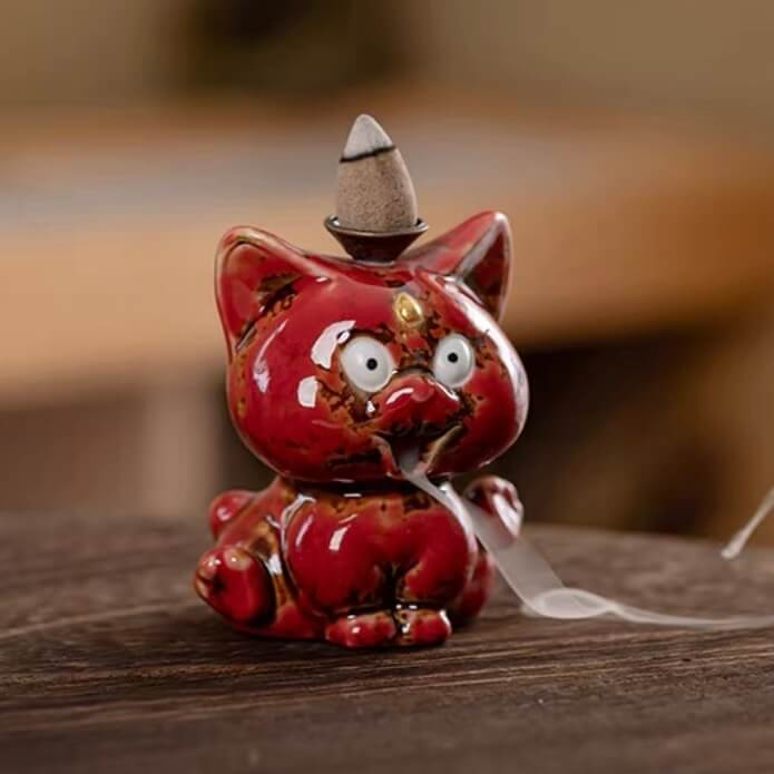 A close-up of a glossy red ceramic cute animal backflow incense burner sitting on a wooden table, featuring a lit incense cone on top with a soft trail of smoke gently flowing downwards from its mouth.