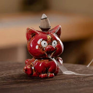 A close-up of a glossy red ceramic cute animal backflow incense burner sitting on a wooden table, featuring a lit incense cone on top with a soft trail of smoke gently flowing downwards from its mouth.