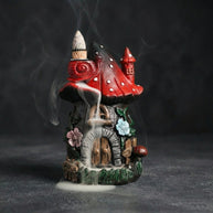 Resin mushroom backflow incense burner shaped like a red mushroom house with cascading smoke, floral details, and a fairy cottage design on a dark background