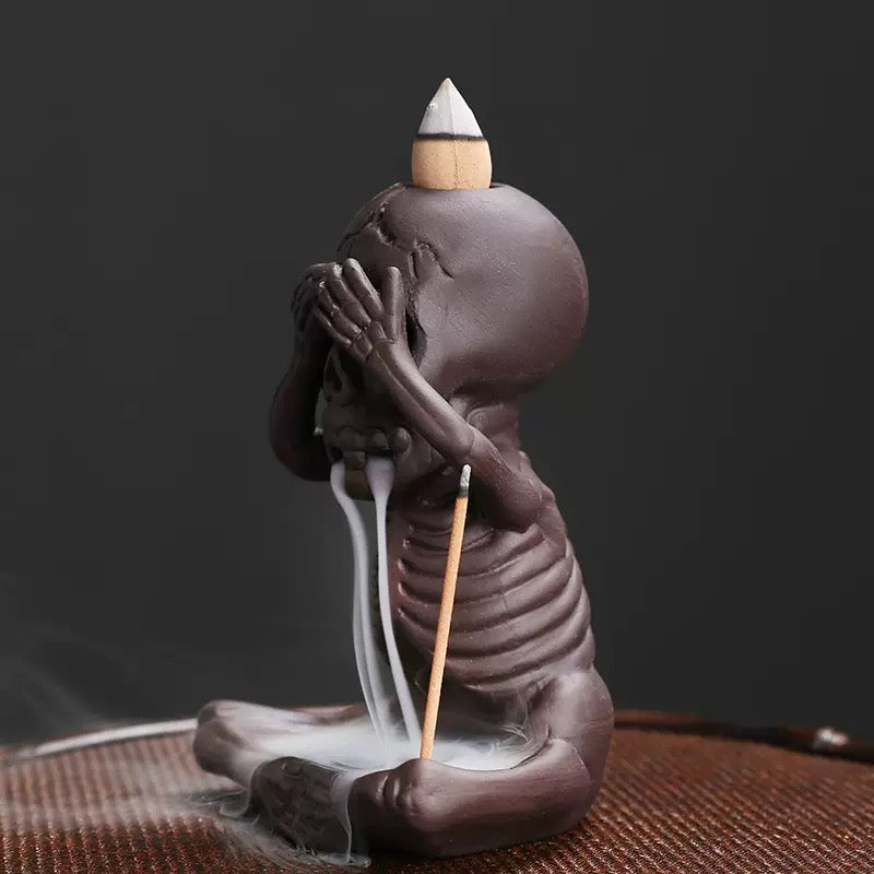 Side view of a seated skeleton backflow incense burner in a “see no evil” pose, with a cone incense on top of the skull, white smoke flowing from the mouth into the base, and a stick incense holder at the side.