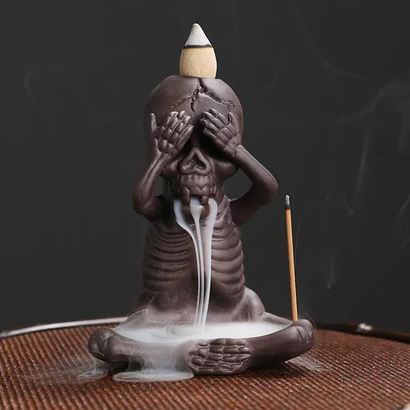  A seated skeleton incense burner covers its eyes in a “see no evil” pose, with a backflow cone on its head releasing white cascading smoke into the base dish and a slim stick incense holder at the side.