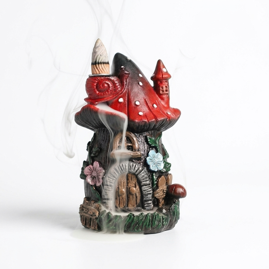 Front view of the whimsical red mushroom fairy house backflow incense burner on a bright white background. Displays the soothing smoke waterfall effect cascading from the snail on the red spotted roof down over the intricate stone doorway and colorful floral details. Perfect for relaxing home decor.