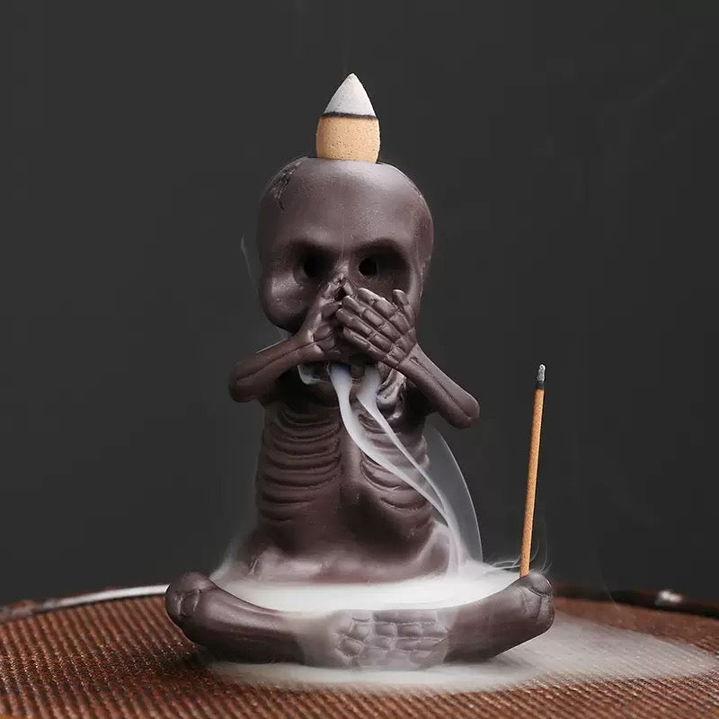 A single seated skeleton incense burner covers its mouth in a “speak no evil” pose, with a backflow cone on its head releasing white cascading smoke and a stick incense holder positioned at the side.