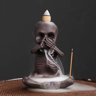 A single seated skeleton incense burner covers its mouth in a “speak no evil” pose, with a backflow cone on its head releasing white cascading smoke and a stick incense holder positioned at the side.