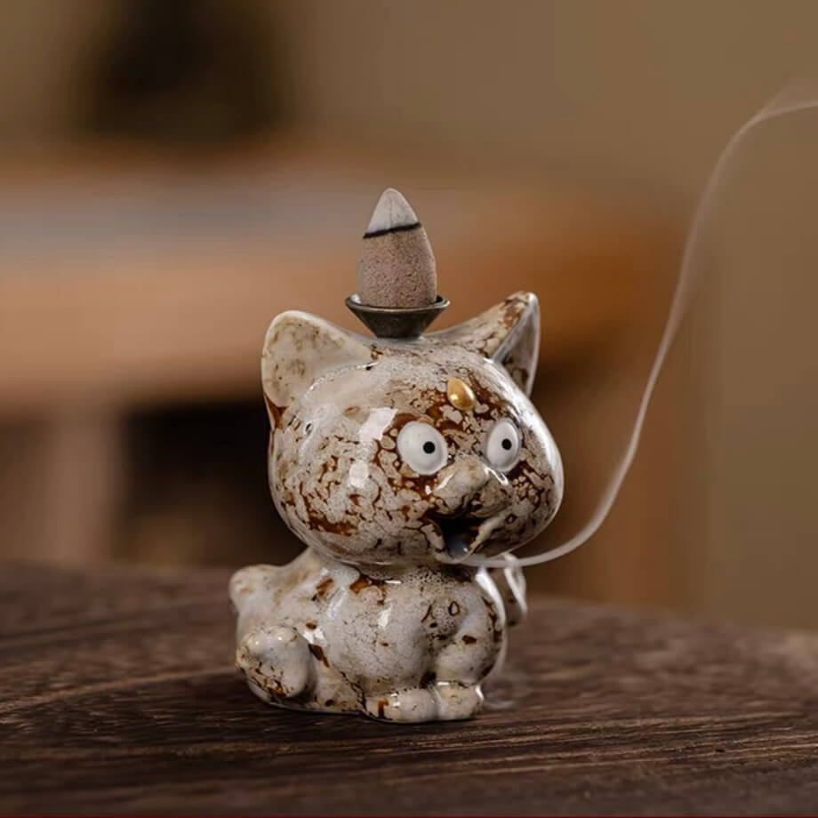 A close-up of a speckled beige and brown ceramic cute animal backflow incense burner sitting on a wooden surface, with a lit incense cone on top and a delicate trail of smoke flowing downwards.