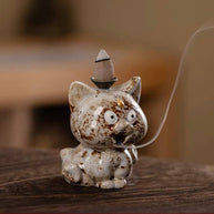 A close-up of a speckled beige and brown ceramic cute animal backflow incense burner sitting on a wooden surface, with a lit incense cone on top and a delicate trail of smoke flowing downwards.