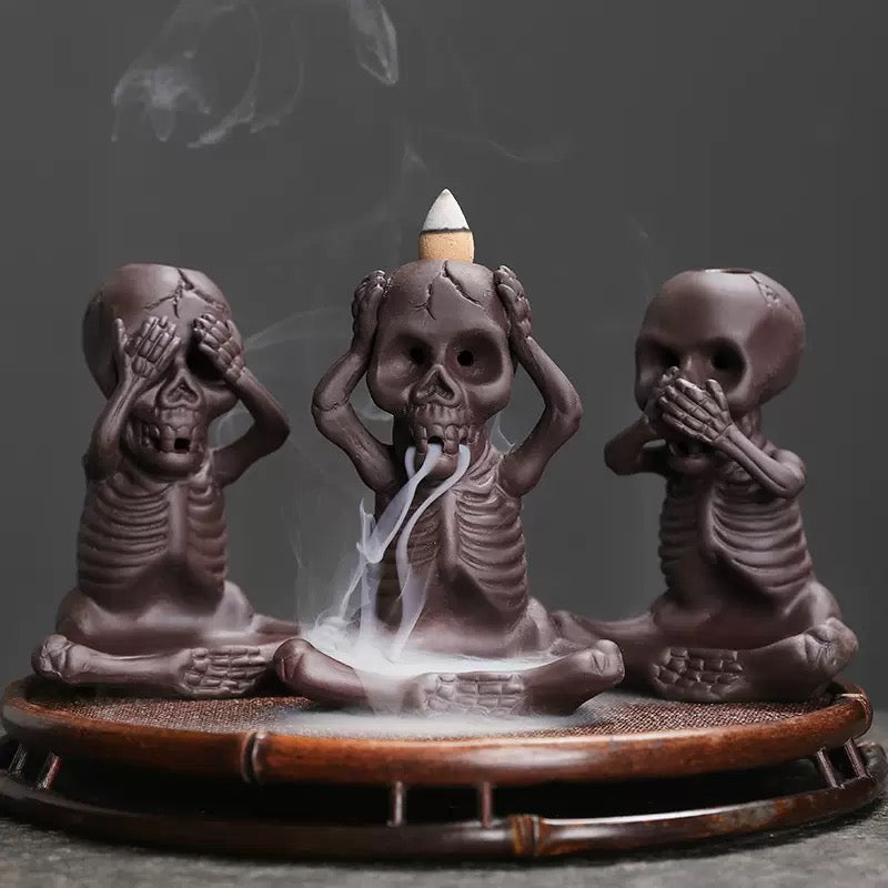 Three small skeleton incense burners in see-no-evil, hear-no-evil, and speak-no-evil poses sit on a round bamboo-style tray, with smoke flowing from the center skull beneath a lit backflow cone.