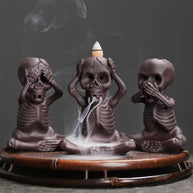Three small skeleton incense burners in see-no-evil, hear-no-evil, and speak-no-evil poses sit on a round bamboo-style tray, with smoke flowing from the center skull beneath a lit backflow cone.