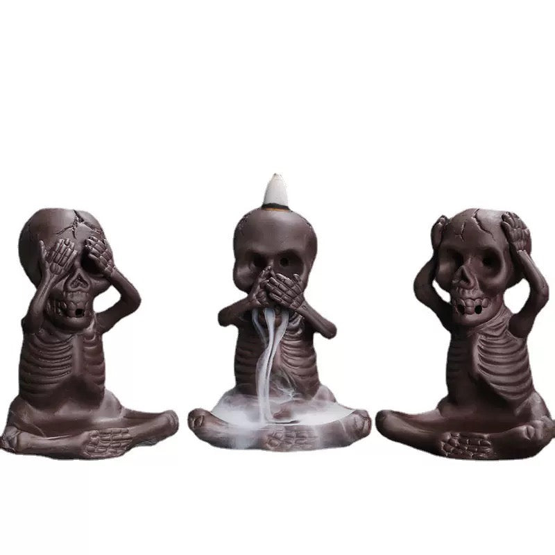 Set of three dark ceramic skeleton backflow incense burners in “see no evil, speak no evil, hear no evil” poses, with white smoke flowing from the center figure onto the base.