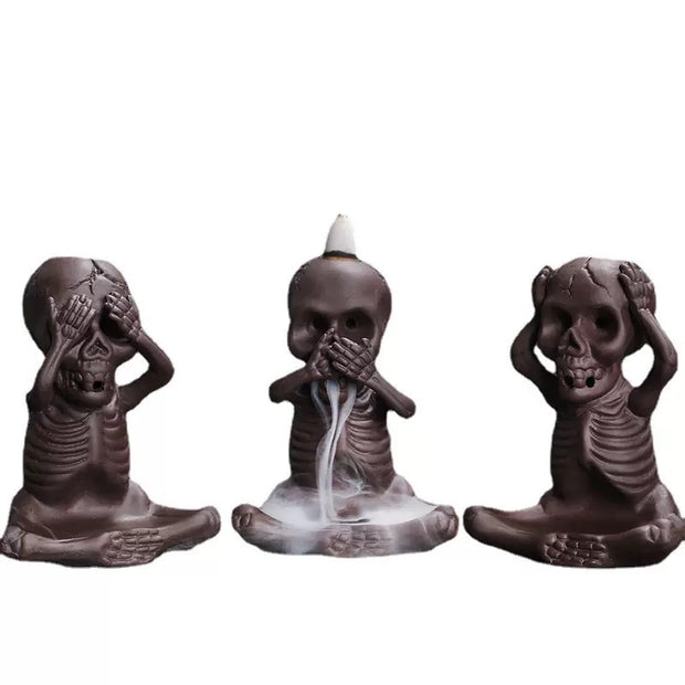 Set of three dark ceramic skeleton backflow incense burners in “see no evil, speak no evil, hear no evil” poses, with white smoke flowing from the center figure onto the base.