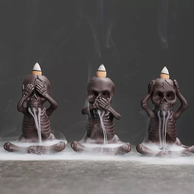 Three seated skeleton backflow incense burners arranged side by side with lit incense cones on their heads, releasing white smoke that streams downward in dramatic trails against a dark background.
