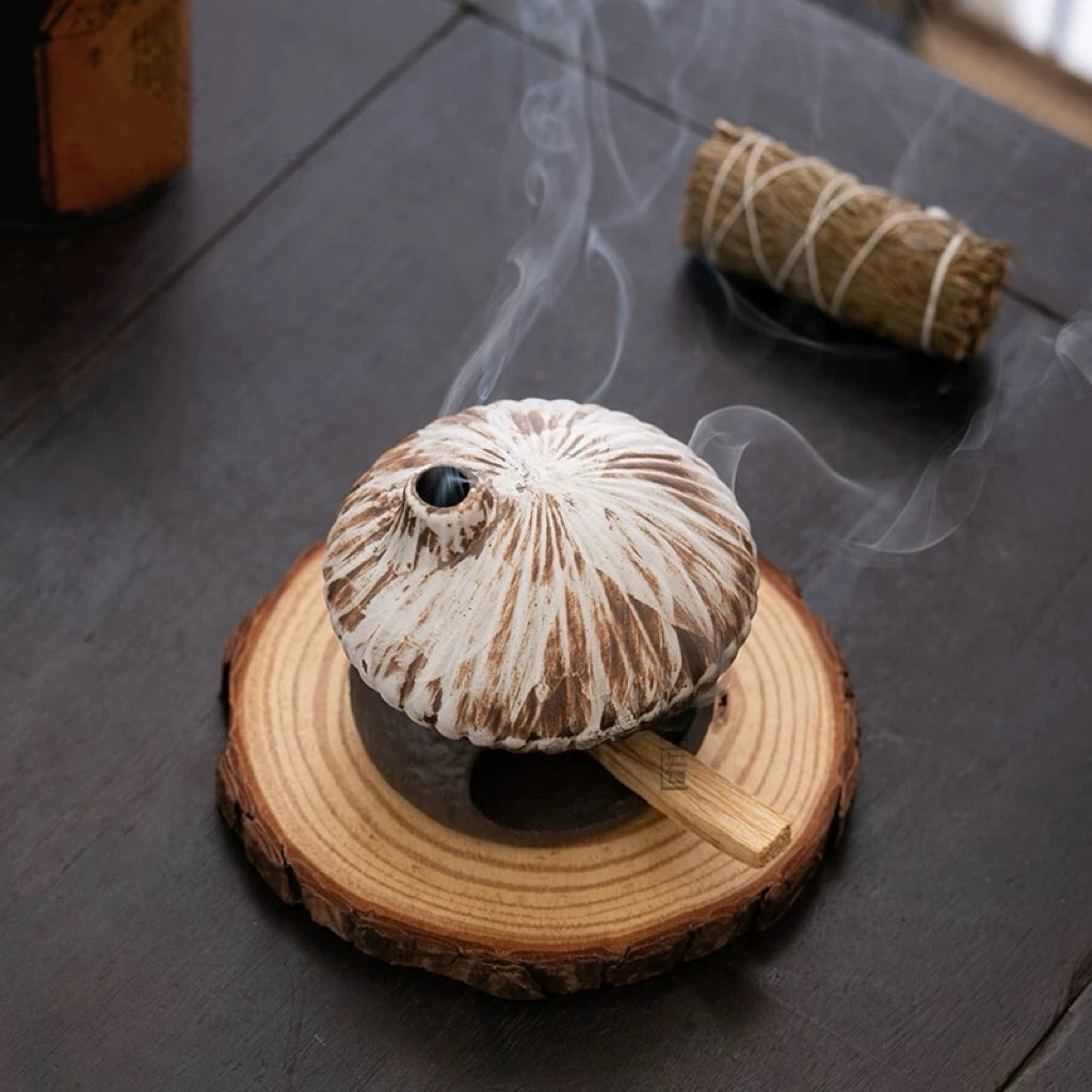 High angle view of rustic ceramic mushroom house incense burner sitting on a wood slice coaster with smoke rising from the chimney and a sage bundle in the background