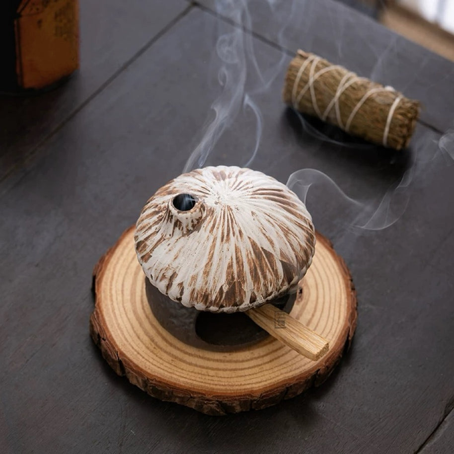 High angle view of rustic ceramic mushroom house incense burner sitting on a wood slice coaster with smoke rising from the chimney and a sage bundle in the background