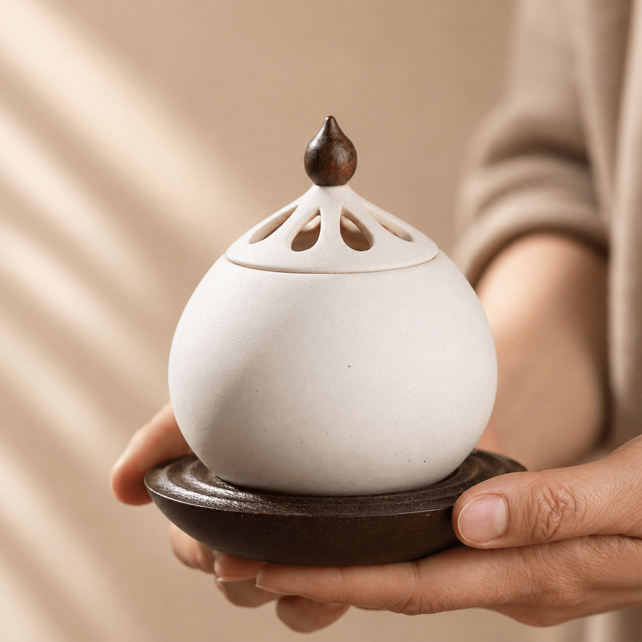 Two hands gently holding a white electric ceramic incense burner by its solid wood base.