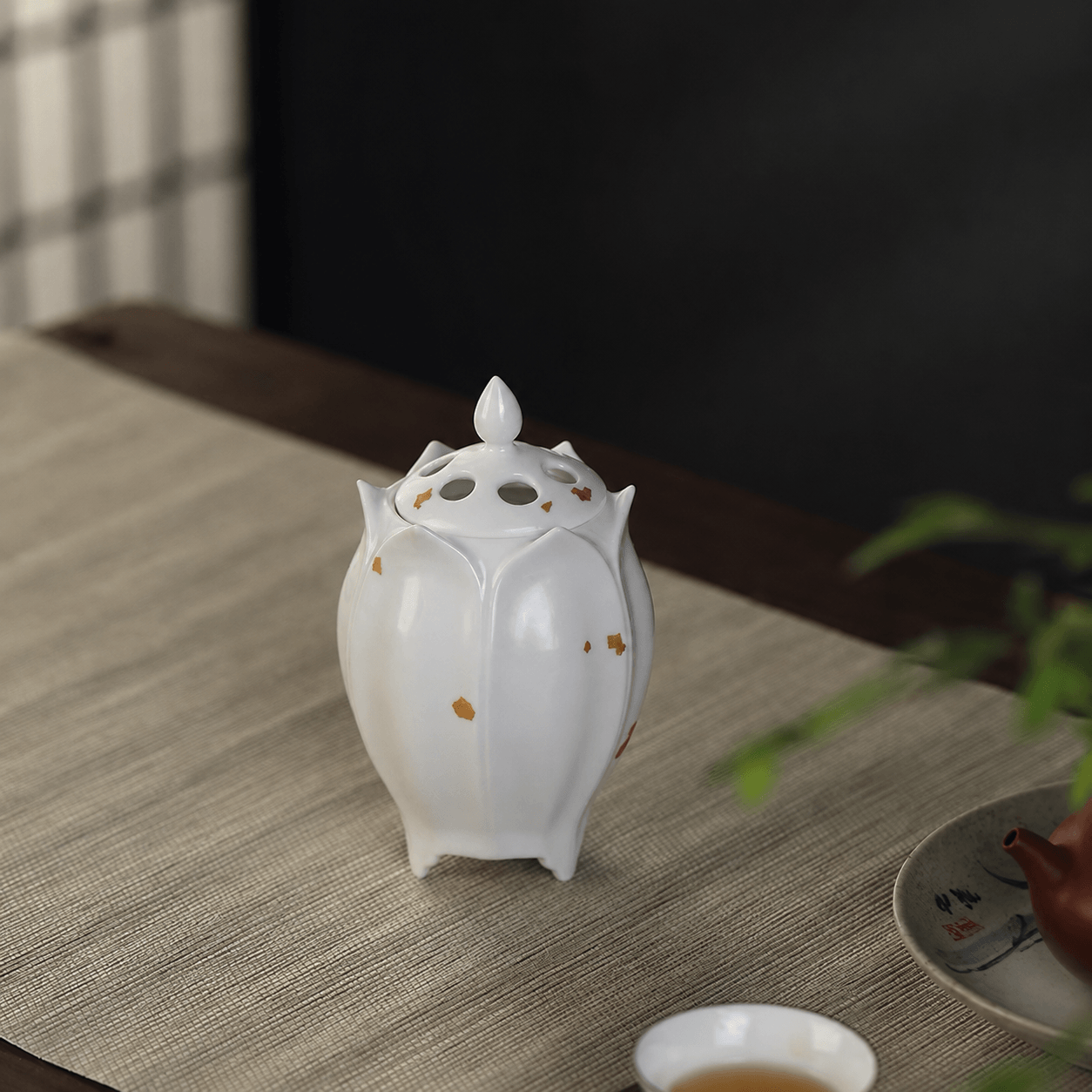 White lotus shaped electric ceramic incense burner with gold flecks displayed on a tabletop beside a tea cup and teapot