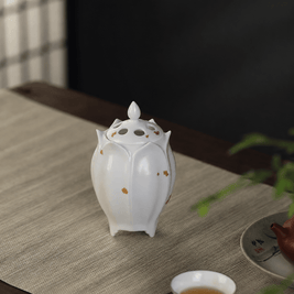 White lotus shaped electric ceramic incense burner with gold flecks displayed on a tabletop beside a tea cup and teapot
