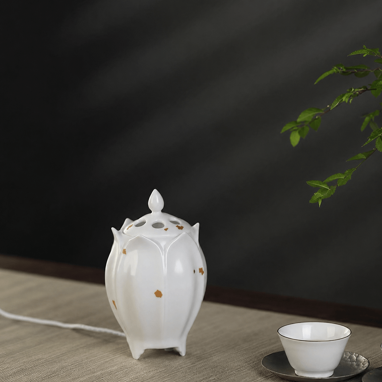 White lotus shaped electric ceramic incense burner with gold flecks displayed beside a tea cup on a light wooden table