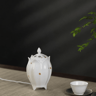 White lotus shaped electric ceramic incense burner with gold flecks displayed beside a tea cup on a light wooden table
