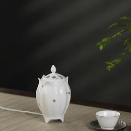 White lotus shaped electric ceramic incense burner with gold flecks displayed beside a tea cup on a light wooden table