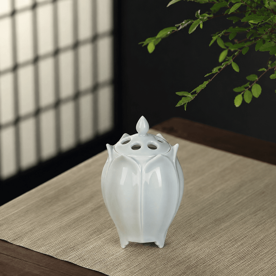 White lotus shaped electric ceramic incense burner displayed on a light wooden table beneath green branches