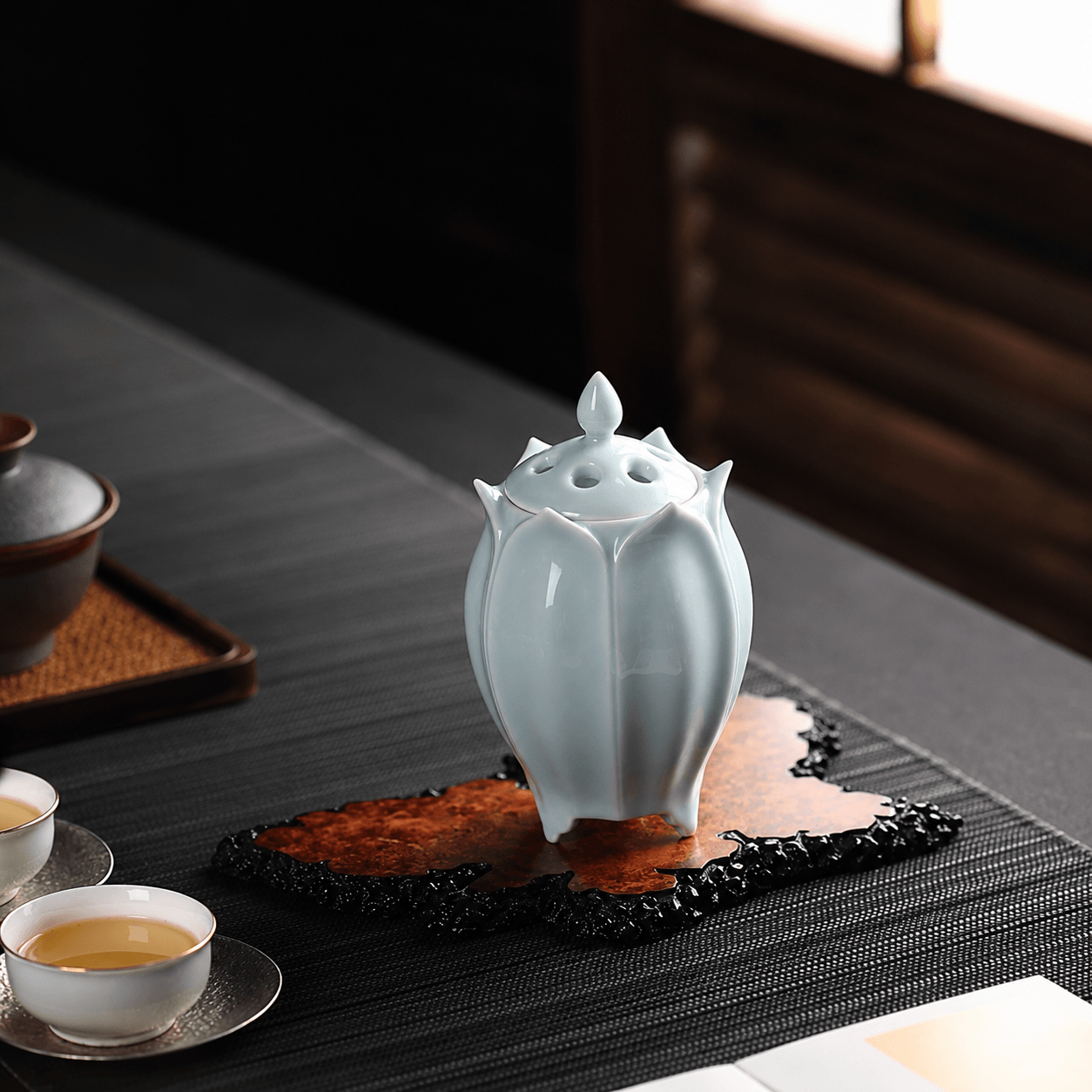 White lotus shaped electric ceramic incense burner displayed on a decorative tray beside tea cups in a sunlit tea room