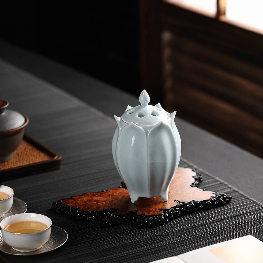 White lotus shaped electric ceramic incense burner displayed on a decorative tray beside tea cups in a sunlit tea room