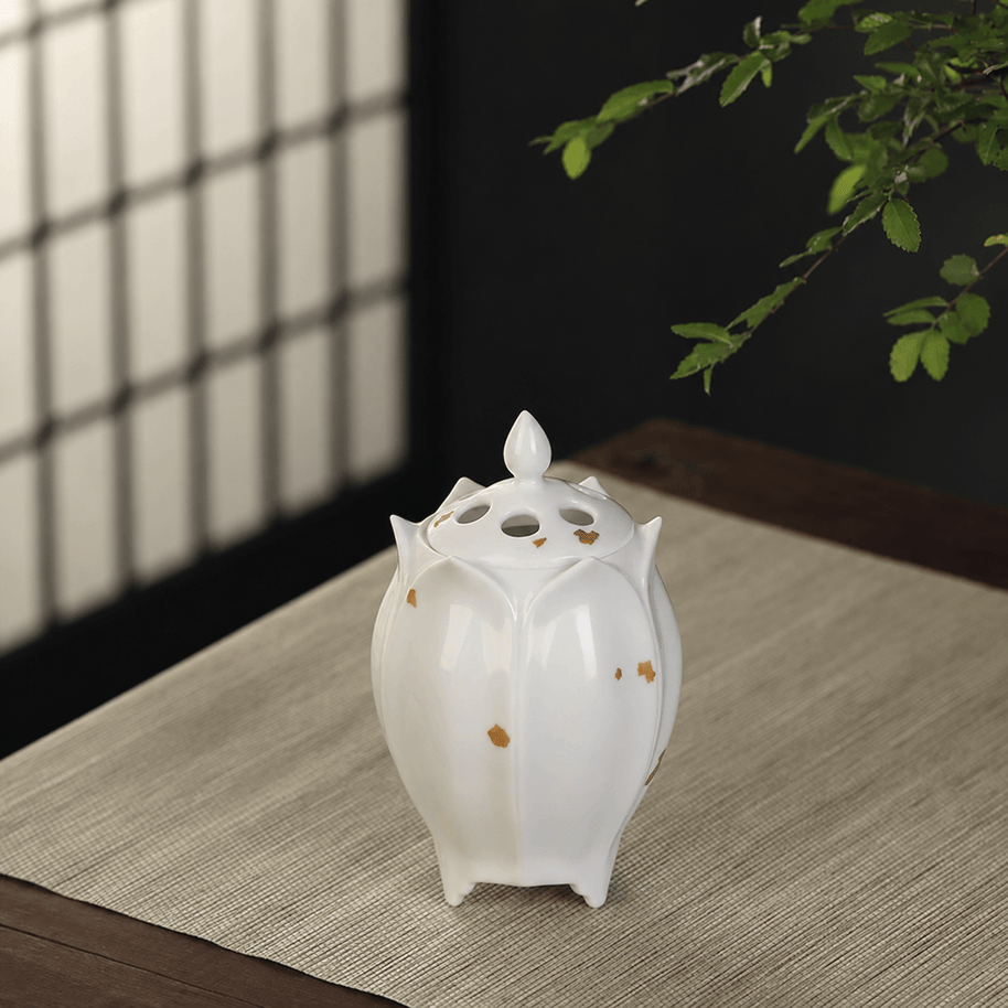 White lotus shaped electric ceramic incense burner with gold flecks displayed on a light wooden table beneath green leaves