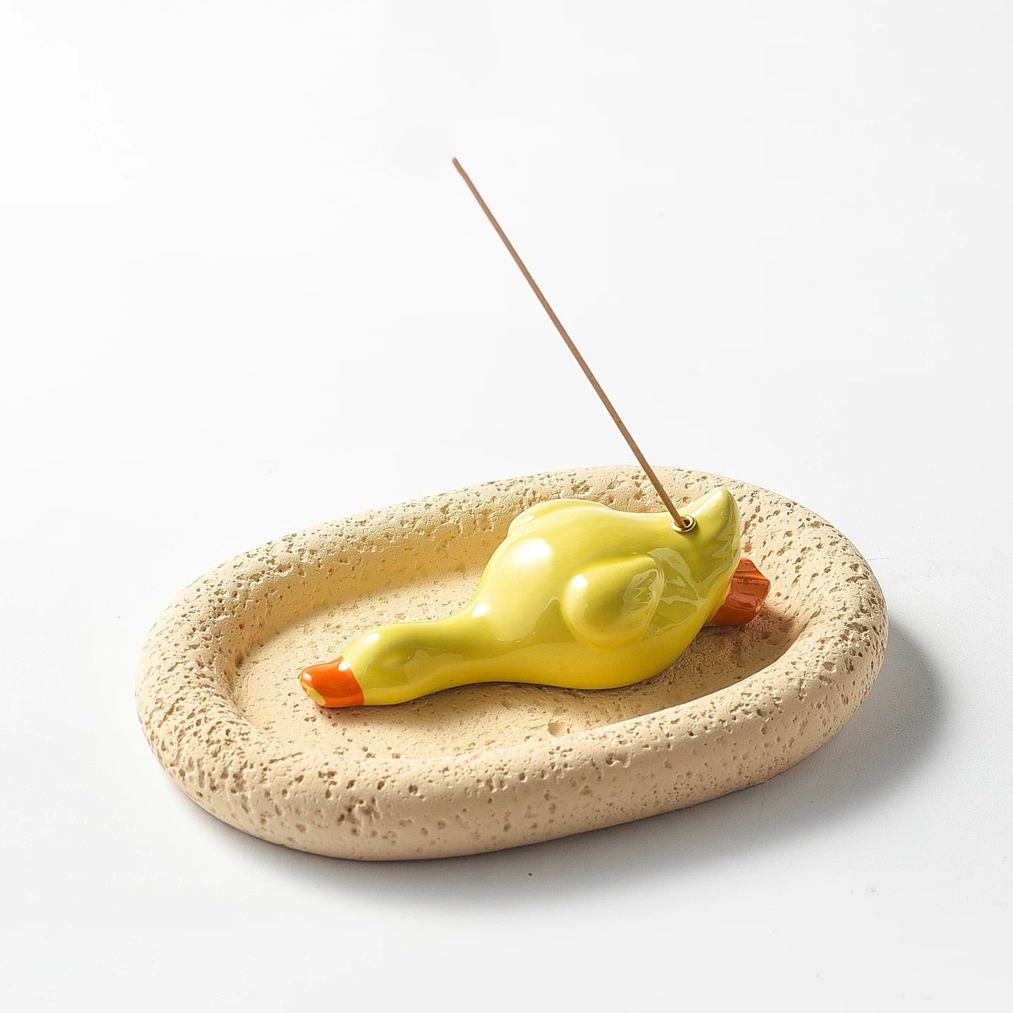 A yellow ceramic duck incense holder resting on a porous beige oval tray. An incense stick is inserted into the duck's back, demonstrating the assembled set in use against a white background.