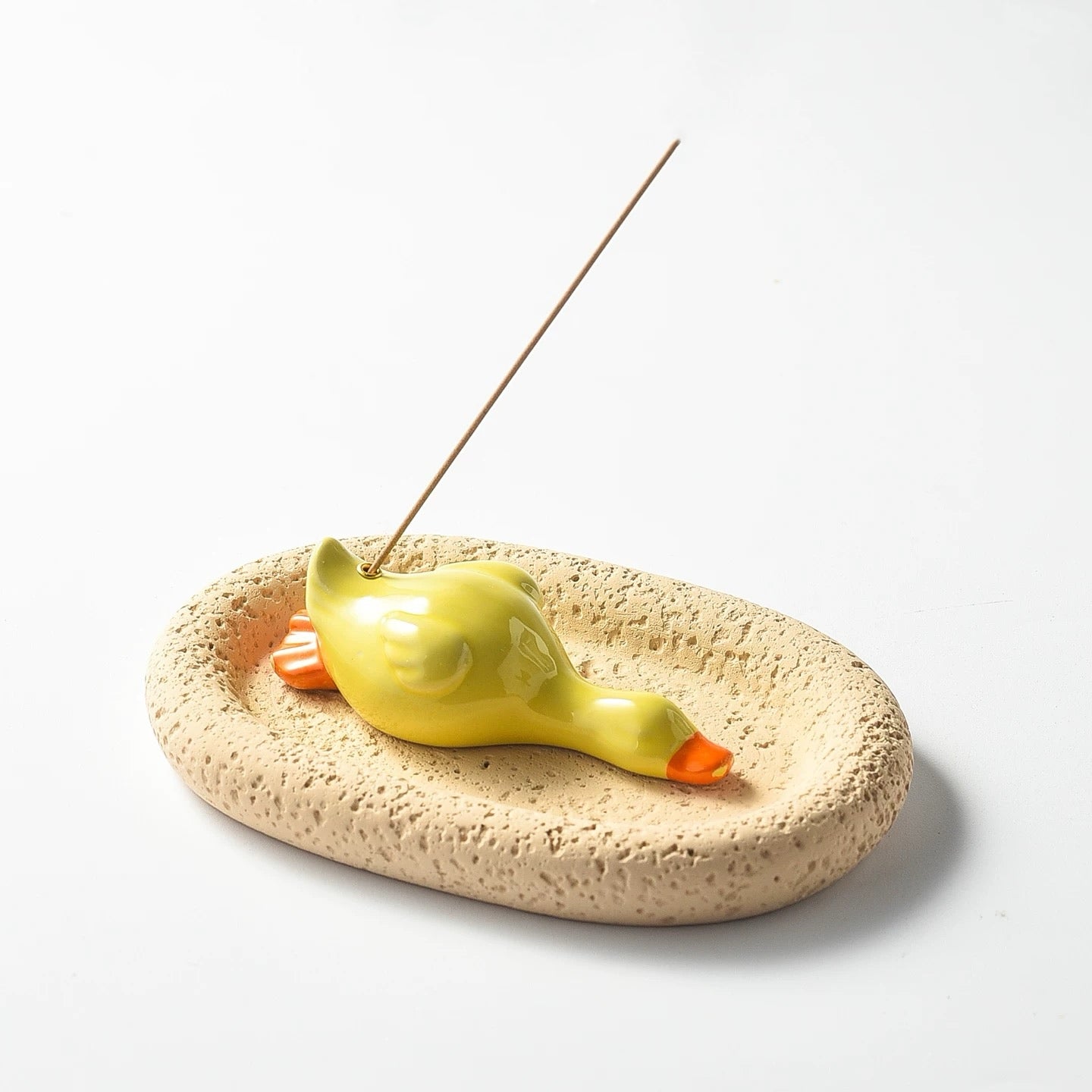 Rear-side view of a yellow ceramic duck incense holder resting on a porous beige tray. An incense stick stands in the holder, and the duck's orange webbed feet are visible from this angle.