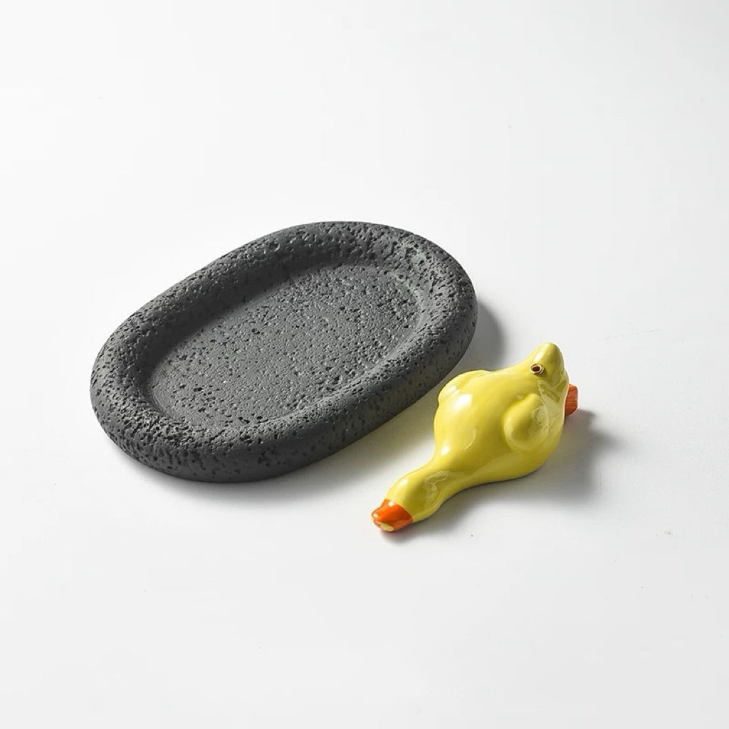 A glossy yellow ceramic duck incense holder paired with a black, porous oval tray. The dark tray provides a strong visual contrast to the bright yellow duck.