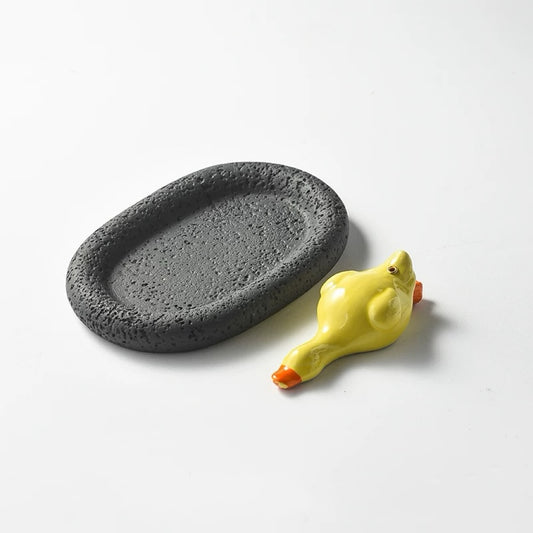 A glossy yellow ceramic duck incense holder paired with a black, porous oval tray. The dark tray provides a strong visual contrast to the bright yellow duck.