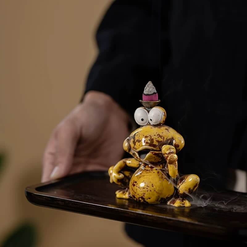 A person holding a dark wooden tray featuring a glossy yellow speckled ceramic backflow incense burner shaped like a frog, with a lit incense cone on its head and smoke beginning to flow downwards.