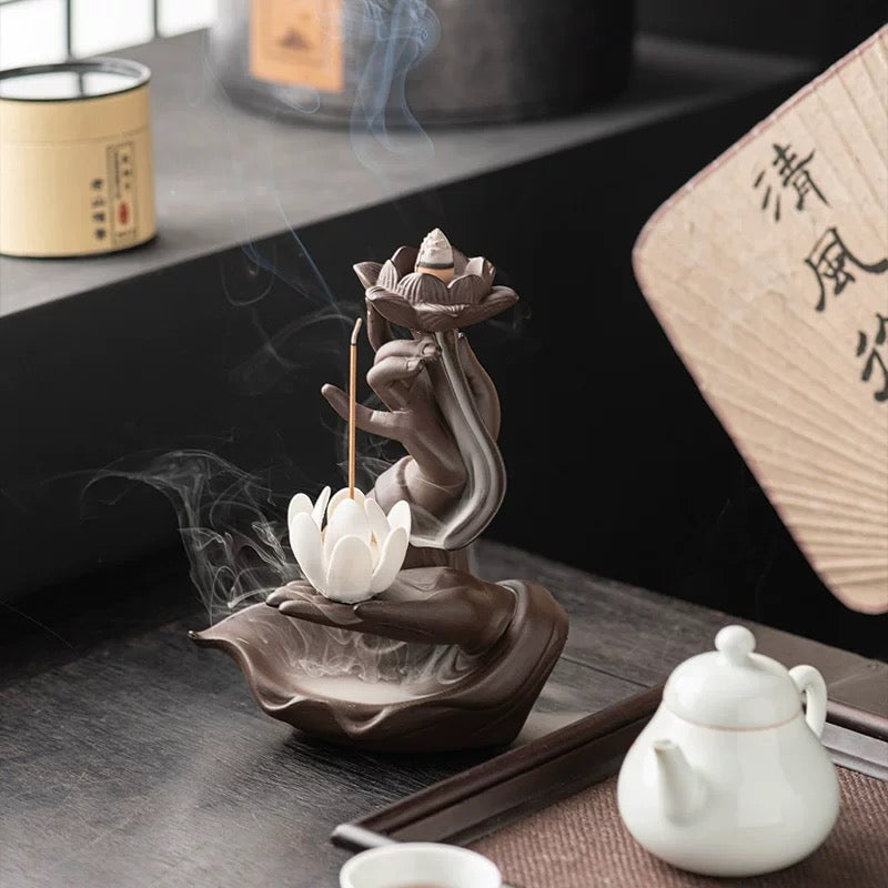 Lifestyle image of a brown hand-shaped lotus backflow incense burner on a tea table, with a lit cone in the upper lotus, a stick incense holder, flowing white smoke, and a white teapot and teacup beside it.