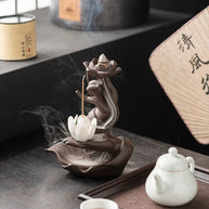 Lifestyle image of a brown hand-shaped lotus backflow incense burner on a tea table, with a lit cone in the upper lotus, a stick incense holder, flowing white smoke, and a white teapot and teacup beside it.