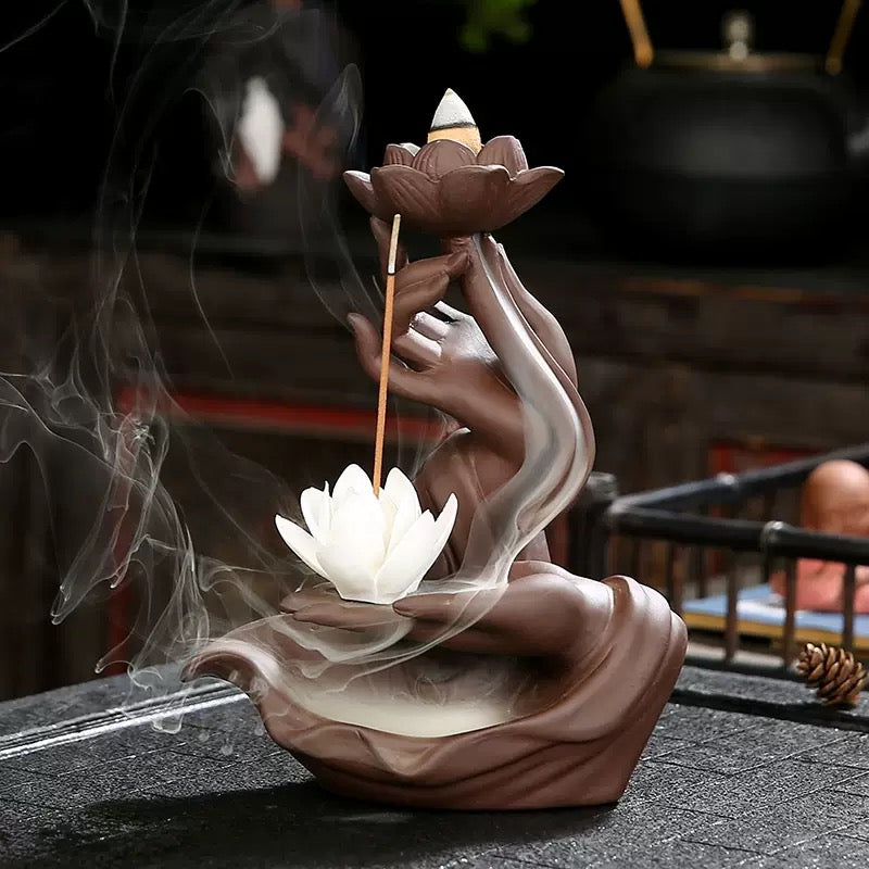 Pink hand-shaped lotus backflow incense burner displayed on a tabletop, with a lit cone resting in the upper lotus, a vertical incense stick beside it, a white lotus below, and soft smoke drifting around the sculpture.