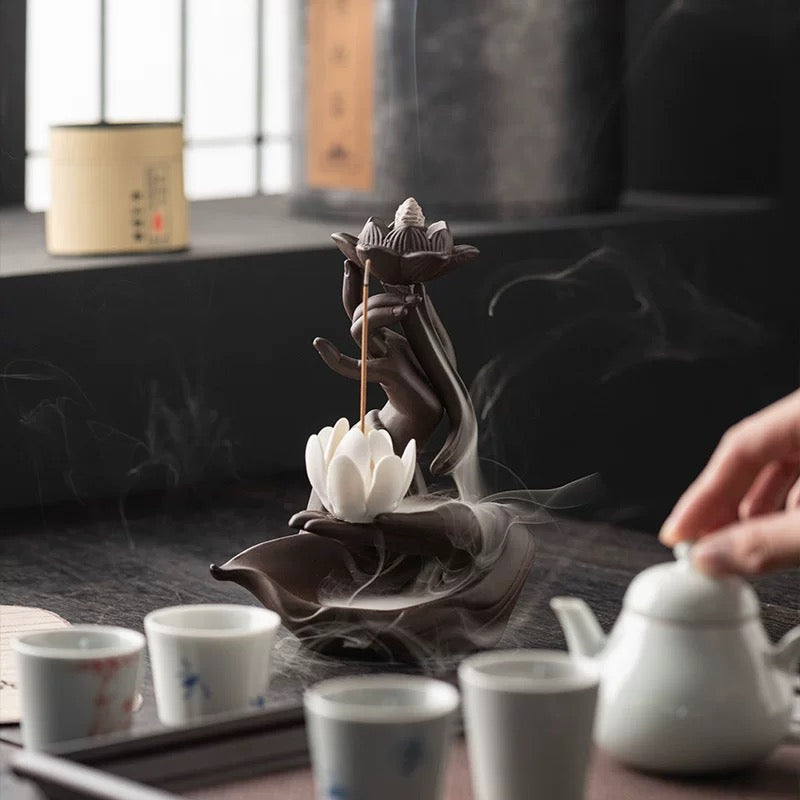 Brown hand-and-lotus backflow incense burner on a tea table, featuring a lit cone in the upper lotus, a white lotus below, curling smoke around the base, and several teacups with a white teapot in the foreground.