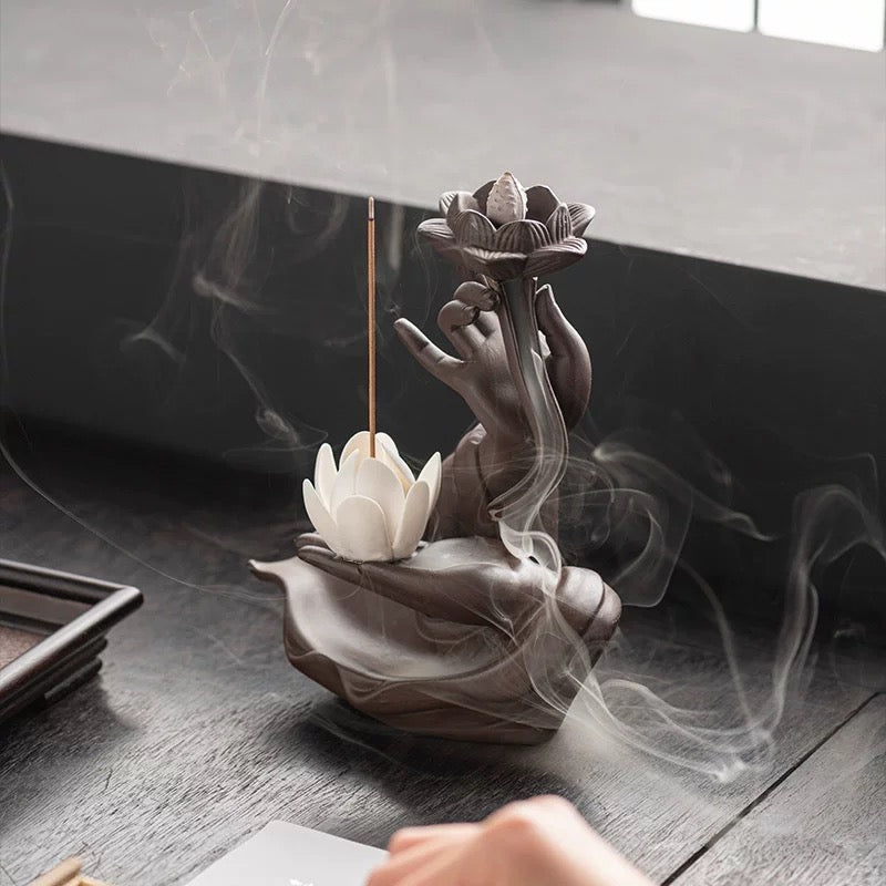 Brown hand-shaped backflow incense burner with a lotus cone holder at the top and a white lotus incense holder below, displayed on a dark tabletop with soft cascading smoke curling around the base.