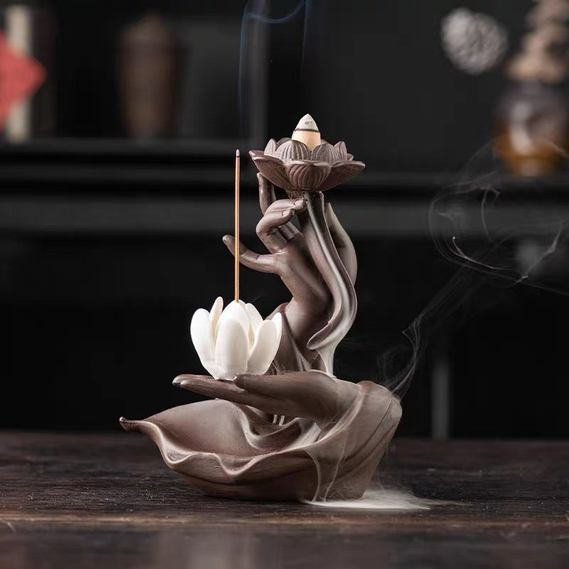 Front-facing product image of a pink hand-shaped lotus backflow incense burner on a wooden surface, featuring a lit cone in the top lotus, a white lotus incense holder below, and soft smoke drifting around the base.