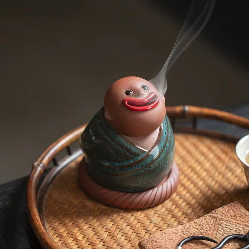 Handmade ceramic incense burner featuring an abstract monk in green robe with red lips, releasing smoke from the mouth while placed on a woven tray