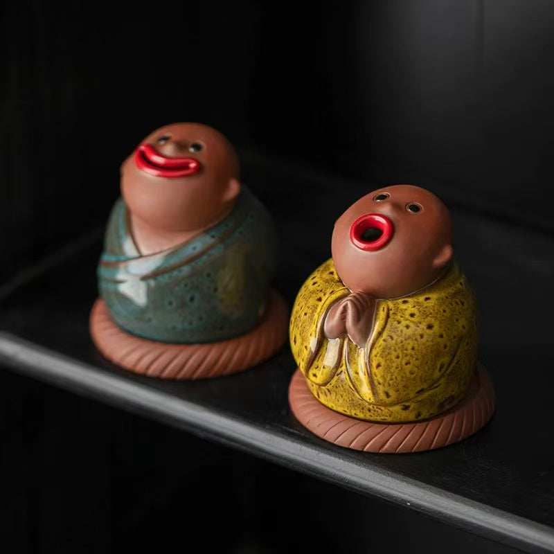 Two abstract ceramic incense burners shaped like monks in yellow and green robes, sitting side by side with open mouths for smoke release