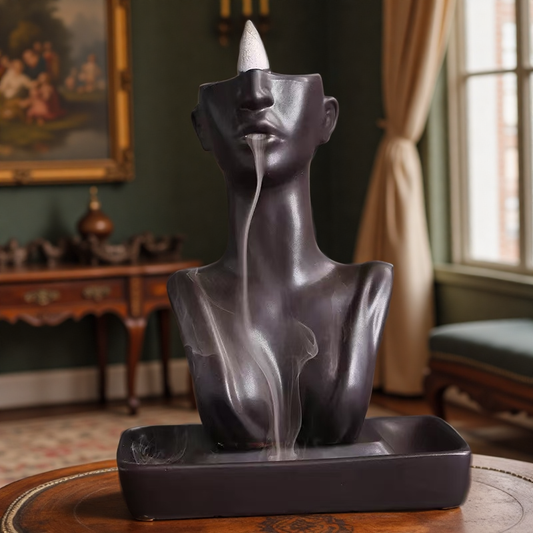 Matte black abstract face backflow incense burner with smoke flowing from lips into rectangular base tray on wooden table