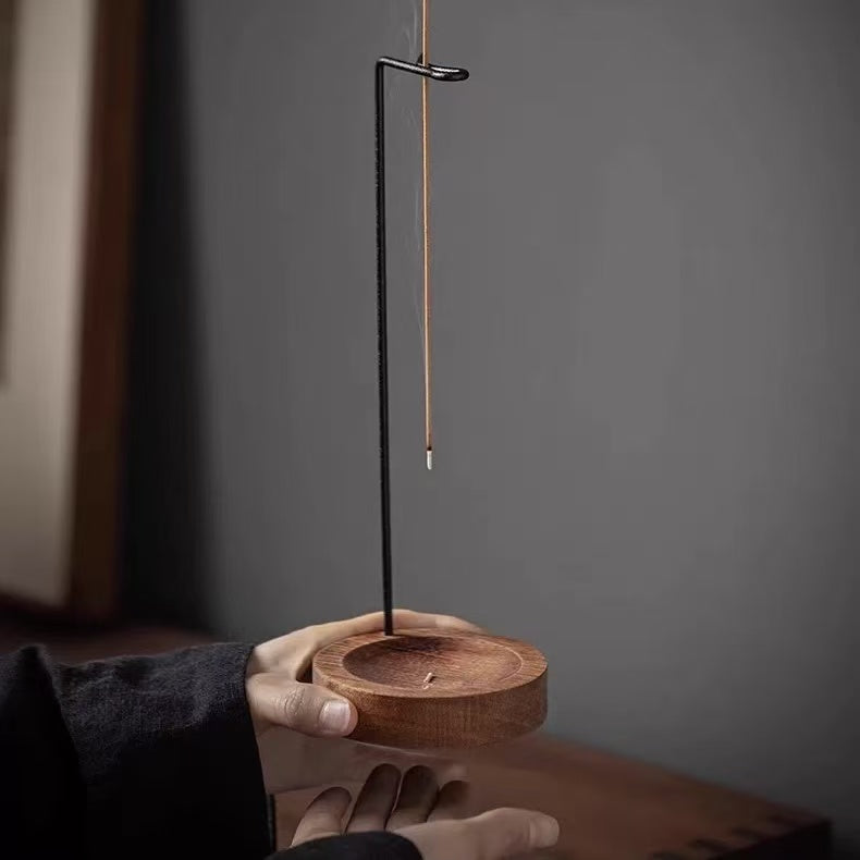 Hand holding a backflow incense burner with wooden base and hanging stick holder, close-up against a neutral wall