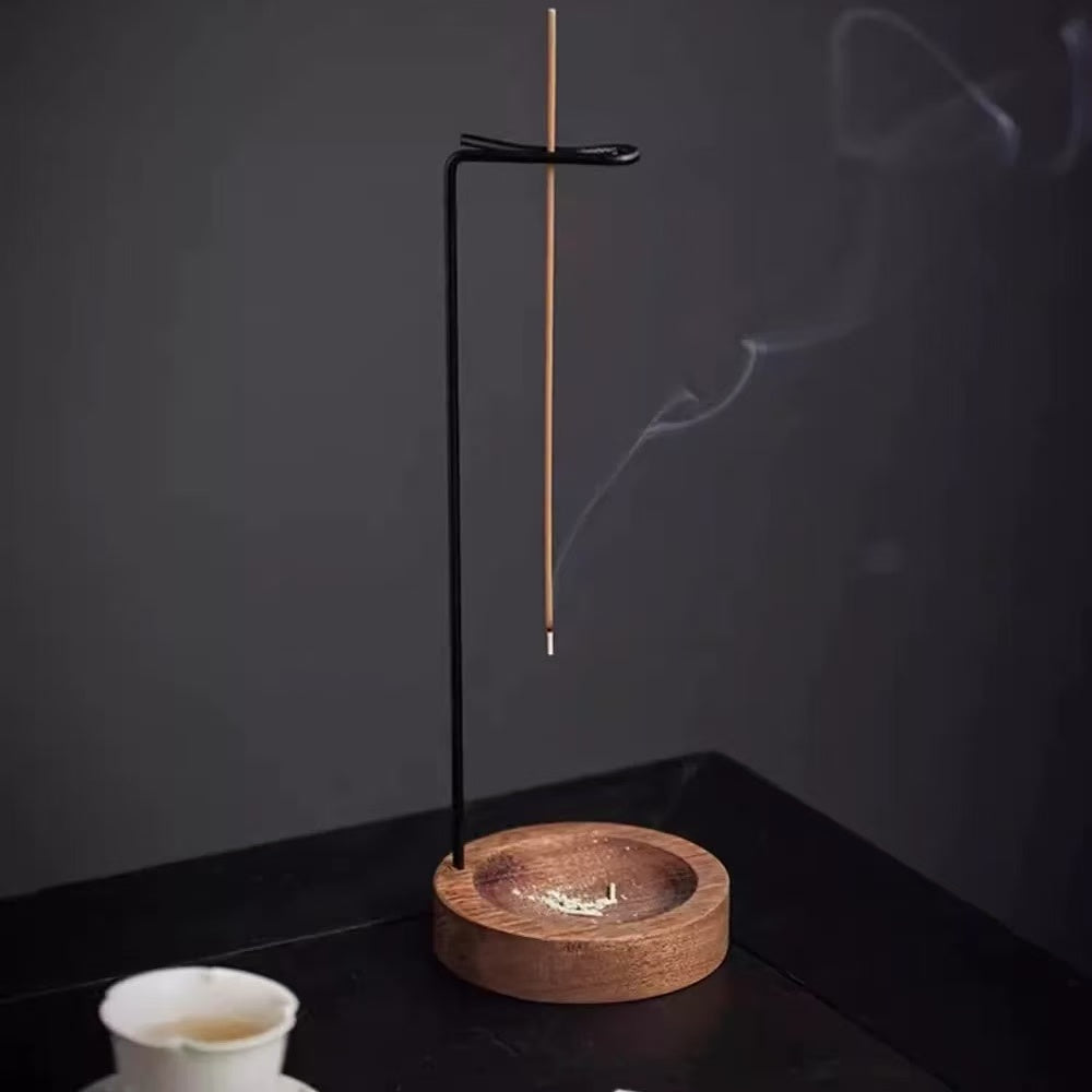 Backflow incense burner with wooden base and hanging stick holder on a dark table, gray backdrop, smoke drifting, small teacup on the left