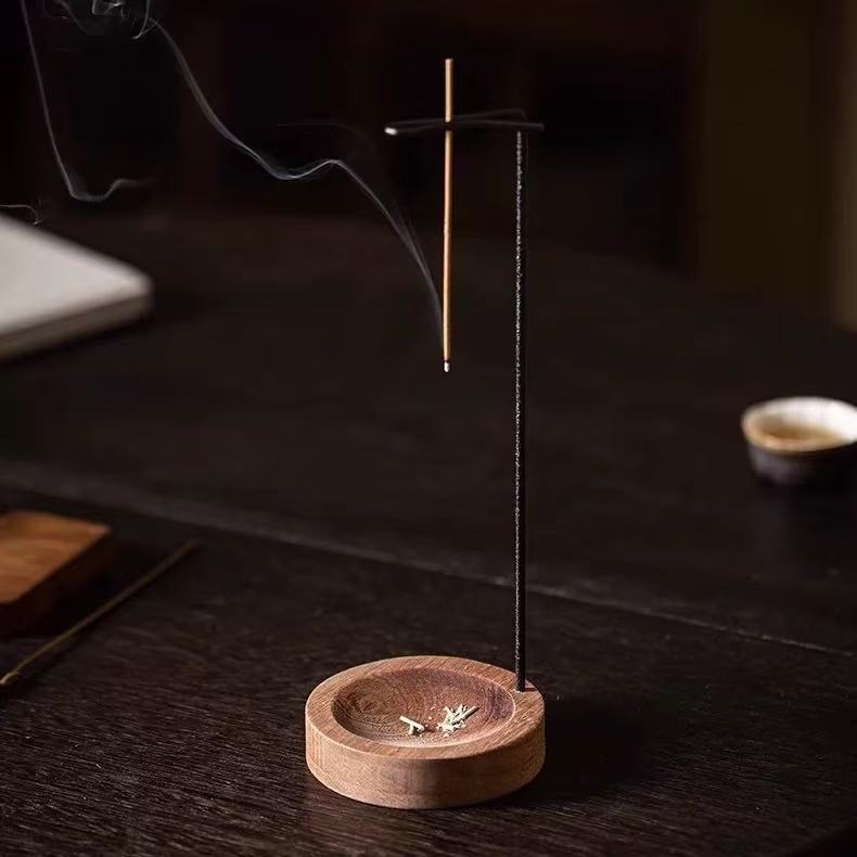 Close-up of a backflow incense burner with wooden base and hanging stick holder on a dark table, ash in the dish and smoke curling, blurred teacup in background