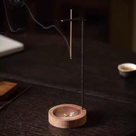 Close-up of a backflow incense burner with wooden base and hanging stick holder on a dark table, ash in the dish and smoke curling, blurred teacup in background
