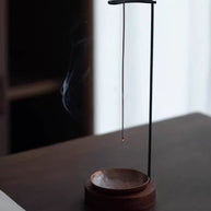 Backflow incense burner with wooden base and hanging stick holder in soft window light on a tabletop, minimal scene, faint smoke