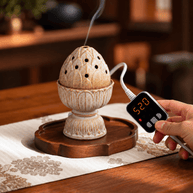 Beige lotus electric incense burner on a wooden tray while a hand holds the digital controller displaying 520°C.
