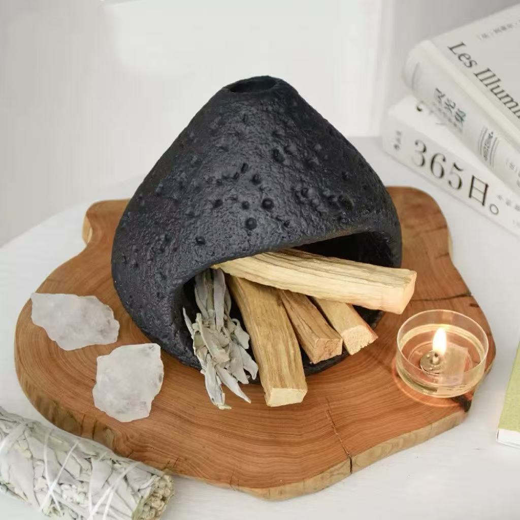 Black natural stone incense burner with palo santo sticks and sage bundle inside, displayed on a carved wooden tray with raw crystals, a burning tealight candle, and books in the background