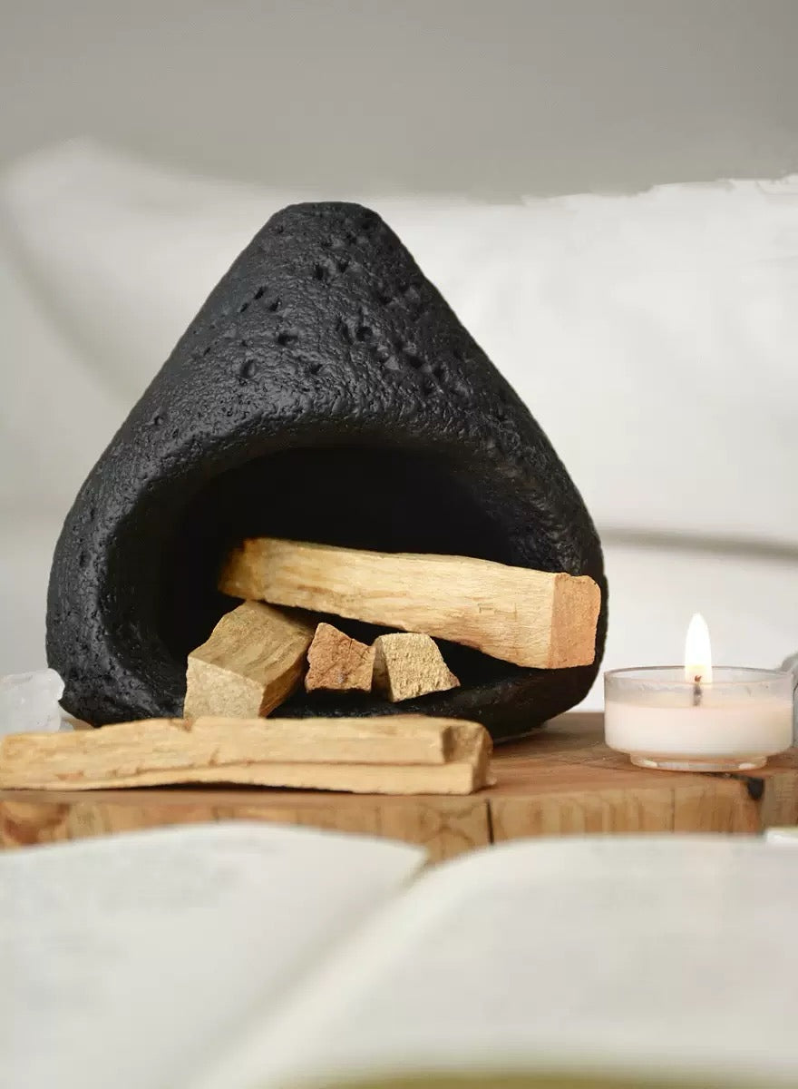 Black natural stone incense burner filled with palo santo wood pieces, displayed on a wooden tray with a lit tealight candle against a soft neutral fabric background